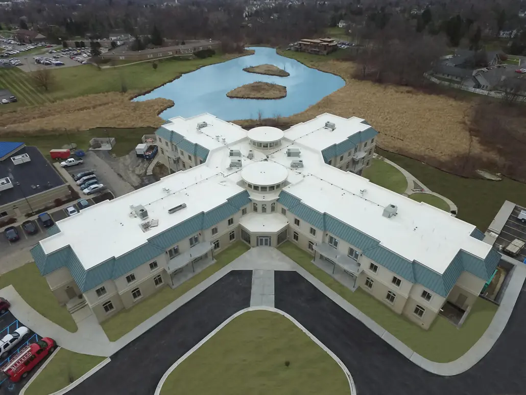 Aerial view of a cross-shaped institutional building with white low-slope membrane roof sections between blue standing-seam metal slopes