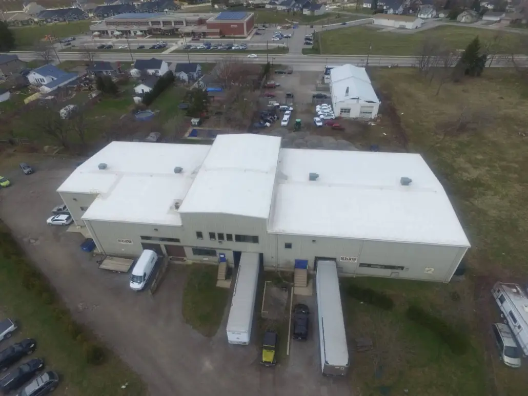 Aerial view of a multi-section commercial building with white low-slope roofs and trailers at the loading doors